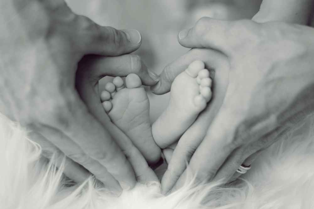 grayscale photo of baby feet with father and mother hands in heart signs
